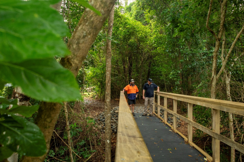 Two men, one wearing hi-vis workwear, inspect a newly built water crossing/bridge in the completed Palm Cove to Ellis Beach section of the Wangetti Trail, with lush green trees in the background.